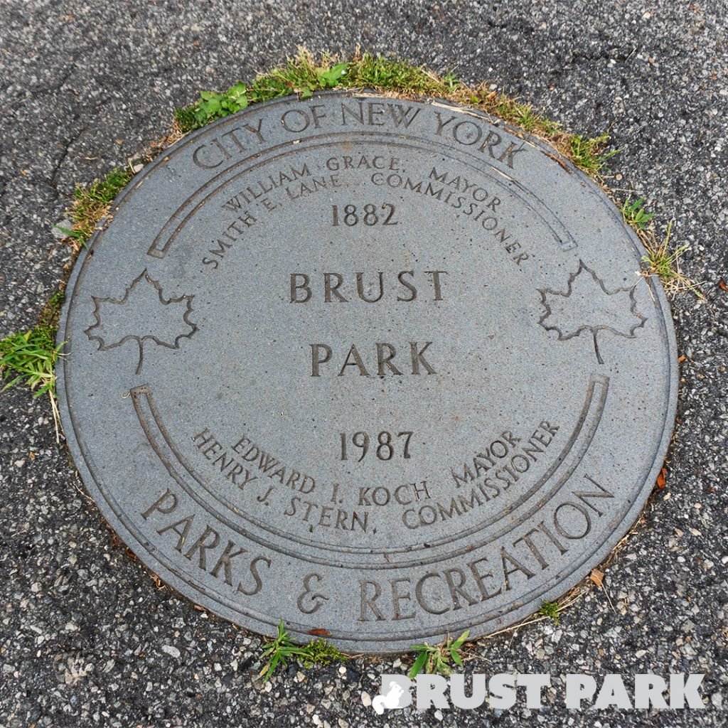 Metal Plate resembling a manhole cover says "Brust Park. City of New York. William Grace, Mayor. Smith E. Lane, Commissioner. 1882. Edward I. Koch, Mayorl. Henry J. Stern, Commissionaler. 1987. Parks & Recreation."